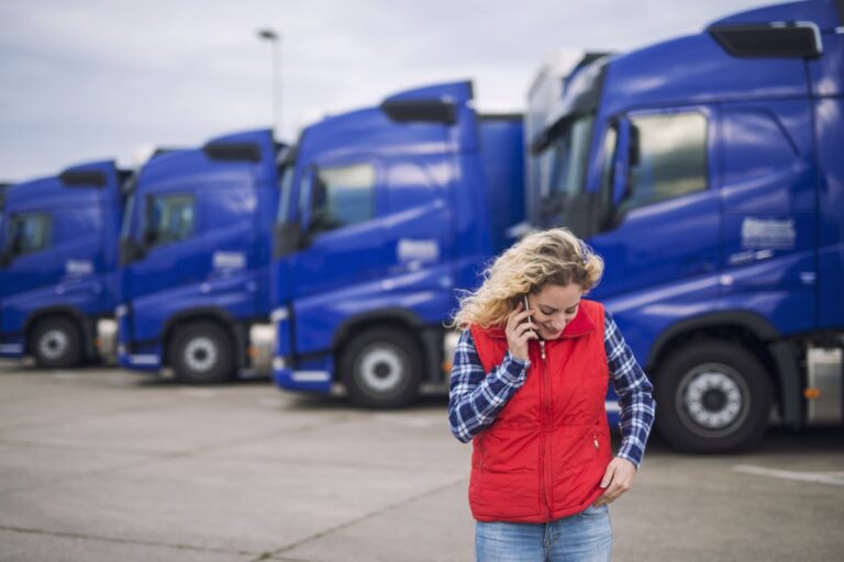 Mujer rubia con indumentaria clásica de camionero hablando por el teléfono móvil, con una flota de camiones azules de fondo.