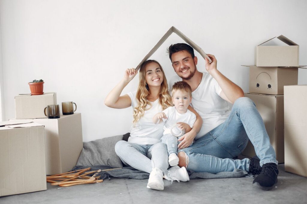 Pareja con niño pequeño entre cajas de mudanza, cubriéndose con un cartón haciendo la forma del tejado de una casa