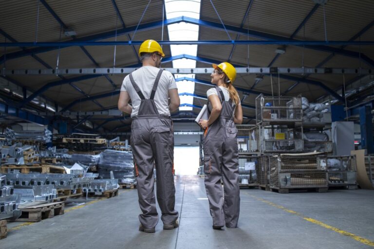 Pareja de obreros (hombre y mujer) con indumentaria de trabajo, de espaldas mientras charlan, en una nave industrial